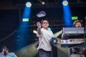 DJ on stage tossing a cap toward the crowd while wearing sunglasses and a white shirt, with blue stage lights in the background and a keyboard rig to the right.