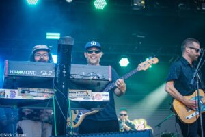 Three musicians on a concert stage: keyboardist on the left, a smiling bassist center wearing sunglasses and a baseball cap, and a guitarist on the right under blue-green stage lights.