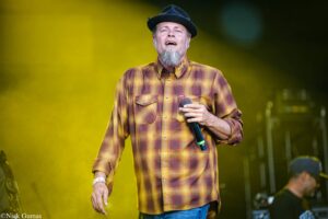 Male singer performing on stage, wearing a yellow plaid shirt and black hat, holding a microphone and singing into the crowd with a yellow-lit backdrop