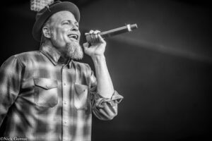 Male singer on stage, wearing a plaid shirt and hat, passionately singing into a handheld microphone.