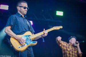 Guitarist wearing sunglasses and a navy patterned shirt plays a Fender electric guitar on stage under colorful lights.