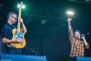 Guitarist in sunglasses plays a yellow electric guitar on stage as a vocalist in a plaid shirt raises a hand and sings nearby.