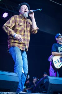 Male singer on stage wearing a brown plaid shirt and a black hat, singing into a microphone with a guitarist visible in the background.