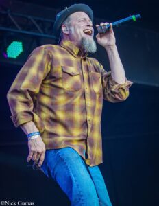 Bearded man in a plaid yellow-brown shirt and hat, singing into a handheld microphone on stage during a performance.