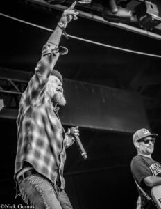Male vocalist on stage, plaid shirt, raising his right arm with index finger pointed upward while singing into a microphone.