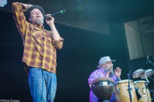 Male singer in a plaid shirt and hat sings into a microphone on stage, one arm raised, performing with a drummer nearby