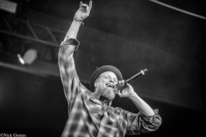 Black-and-white photo of a bearded man in a plaid shirt on stage, raising one arm and singing into a microphone, mid-performance.