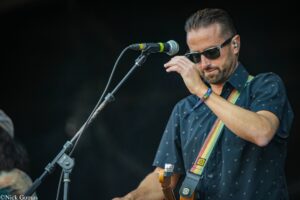 Musician wearing sunglasses singing into a microphone while playing a guitar on stage, dark background behind him.