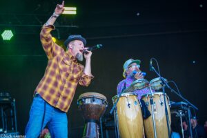 Male singer in a plaid shirt and hat performs into a microphone on stage; a percussionist plays congas beside him under green stage lights.