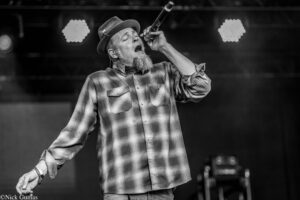 Male singer on stage, wearing a plaid shirt and hat, singing into a handheld microphone under stage lights.