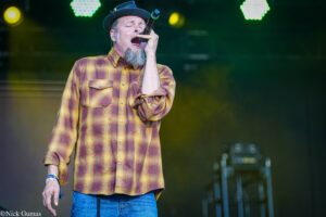 Male singer on stage wearing a yellow plaid shirt and hat, passionately singing into a microphone.