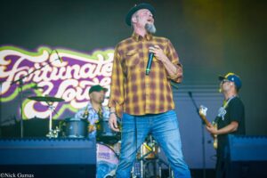 Male singer in a yellow plaid shirt and green hat performs on stage with a microphone, colorful backdrop behind him.