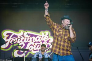 Male singer on stage wearing a yellow plaid shirt and hat, microphone in hand, raising one arm as he performs.
