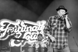 Black-and-white photo of a bearded man in a plaid shirt and hat singing into a microphone on stage with a drummer in the background.