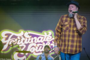 Male singer wearing a brown plaid shirt and black hat, holding a microphone on stage.