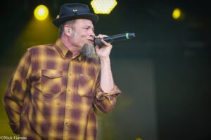 Male singer in a brown plaid shirt and hat, singing into a microphone on stage with yellow lights behind him.