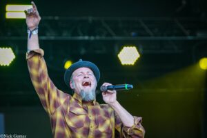 Male singer on stage raising his right arm, holding a microphone, wearing a plaid shirt and blue hat with bright stage lights behind him