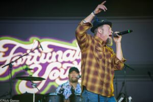 Male singer in a brown plaid shirt and hat performs on stage with a microphone, with a drummer and colorful backdrop behind him.