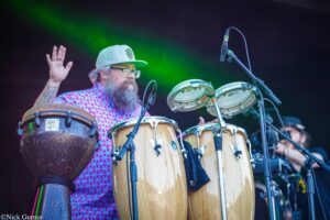 Bearded drummer wearing glasses and a gray cap, playing wooden conga drums on stage with green lights in the background.