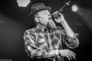 Male singer in a hat and plaid shirt sings into a handheld microphone on stage, eyes closed in passion (black and white).