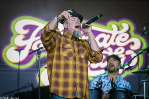 Male singer with a gray goatee wearing a black hat and yellow plaid shirt, singing into a microphone on stage with a colorful mural backdrop.