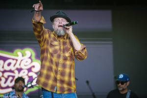 Male singer with a gray beard, wearing a yellow plaid shirt and a hat, performing on stage into a microphone with one arm raised.