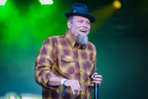 Male musician on stage holding a microphone, wearing a brown plaid shirt and a dark hat, smiling against a green backdrop.