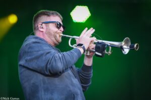 Man in sunglasses playing a trumpet on stage with bright green lights in the background, outdoor concert.