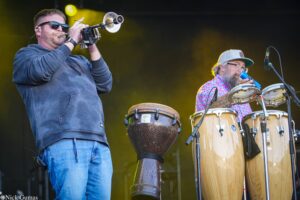 Two musicians performing on stage: a man in a blue hoodie playing a brass trumpet while wearing sunglasses, beside a percussionist with a gray cap playing conga drums.