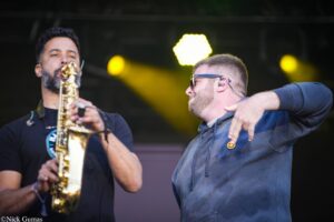 Two male musicians perform on stage: a saxophonist on the left and a man in a blue hoodie on the right, yellow stage lights behind them.