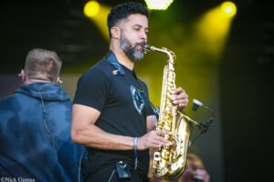 Male saxophonist in a black t-shirt performing on a dimly lit stage with yellow spotlights, playing a gold saxophone; audience blurred in the background.