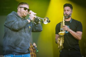 Two musicians perform on a stage: a bearded man in a hoodie playing a silver trumpet, while another with a Saxophone stands nearby under yellow lighting.