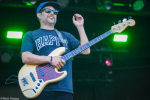 Bassist wearing a blue cap and sunglasses plays a cream-colored bass guitar on a green-lit stage.