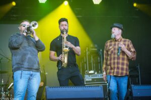 Three musicians perform on a stage under yellow lights: left trumpet player in a hoodie, center saxophonist blowing into his instrument, right singer with a microphone wearing a plaid shirt and hat.
