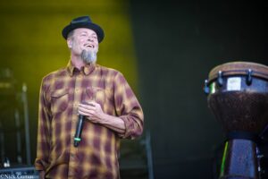 Smiling man with a gray beard and black hat singing into a microphone on stage, wearing a plaid shirt.
