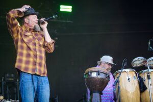 Male singer wearing a plaid shirt and cap performs on stage, holding a microphone near his mouth with a green stage light behind him.