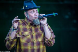 Male singer wearing a blue hat and yellow plaid shirt, singing into a handheld microphone on stage with eyes closed.