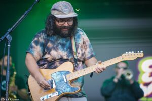 Bearded guitarist in a tie-dye shirt and cap playing a light-colored electric guitar on stage.