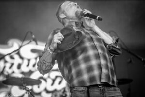 Male singer on stage, singing into a handheld microphone while holding a hat against his chest, wearing a plaid shirt; drums visible in the blurred background.