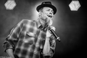 Male singer on stage, wearing a plaid shirt and hat, singing into a handheld microphone in a black-and-white photo.