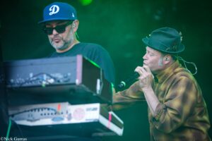 Two male performers on a dimly lit stage: a keyboard/DJ wearing a blue cap and sunglasses, and a singer in a hat with a microphone under green lighting.