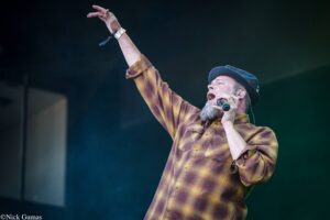 Male singer on stage wearing a plaid shirt and blue hat, raising his right arm while singing into a microphone.