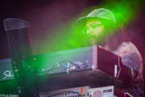 Bearded performer with glasses and a cap using a mixer on a stage with neon green and pink lighting.