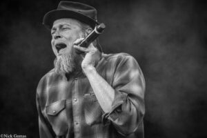 Black-and-white photo of a bearded man wearing a hat and plaid shirt, singing into a handheld microphone on stage.
