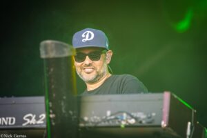 DJ or electronic musician wearing a blue cap with a white 'D' logo and sunglasses, operating a keyboard on a dimly lit stage with green lighting in the background.