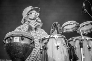 Bearded drummer wearing glasses and a cap plays a drum set with mallets in a black-and-white photo.