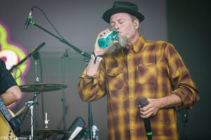 Male musician wearing a brown plaid shirt and black hat drinks water on stage during a performance, microphone in hand nearby