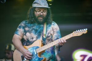 Male guitarist with long hair and beard wearing a tie-dye shirt and cap, playing an electric guitar on stage at a concert.