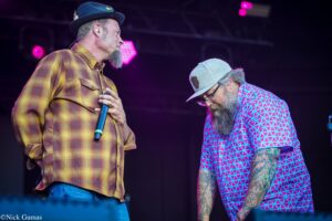 Two male performers on a concert stage: one in a yellow-brown plaid shirt holding a microphone, facing the other who wears a bright geometric shirt and a gray cap.