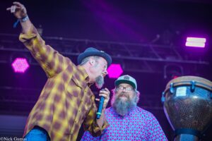 Two male performers on stage singing into microphones under purple stage lights; one wearing a yellow plaid shirt and hat.